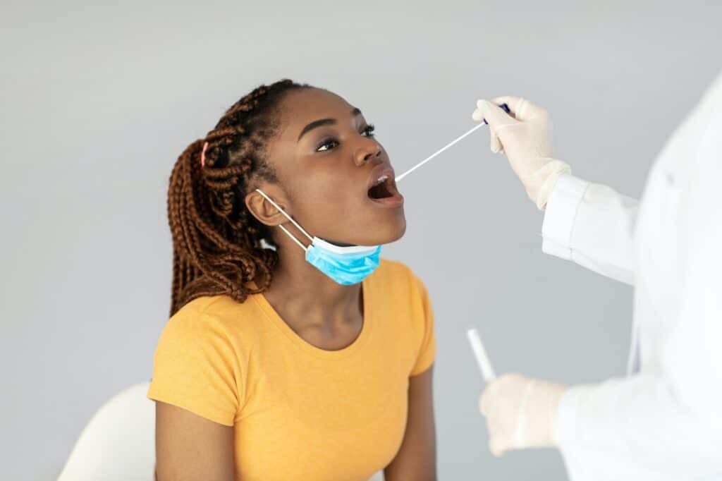 Doctor making oral covid PCR test for young black female patient over grey studio background